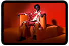 iPad mini displayed, front view, screen showing a vivid screen with a person in an orange suit, sitting on an orange chair, placed in an orange room