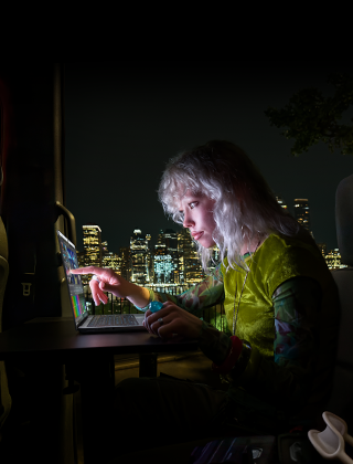 A person sitting in the back seat of a van, the open passenger door reveals a city skyline at night, MacBook Pro with PROLED XDR Display on a worktable, the person is illuminated by the screen while using the Touch feature to edit a video in DaVinci Resolve Studio